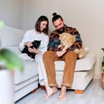 Woman sitting on her living room sofa holding her black cat around her neck and her partner sitting next to her holding a common European cat.