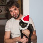 Funny pet owner, man holding cat wearing Santa hat Christmas decoration for holiday season. Cat daddy concept for Santa Claus costume with beard, funny animal clothing, or festive winter holidays. Candid man wearing Santa hat is indoors in domestic room interior at home with his pampered pets. Real life, real people. Part of a series.